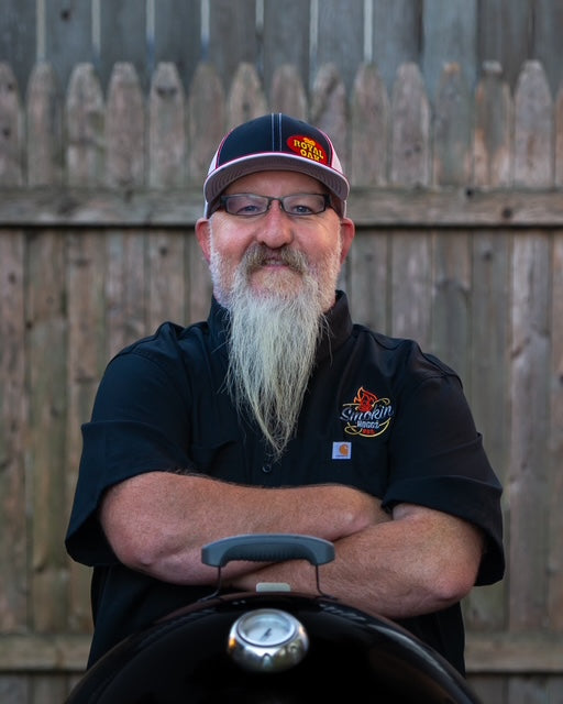Man with a beard and glasses wearing a black shirt with a logo, standing in front of a wooden fence.